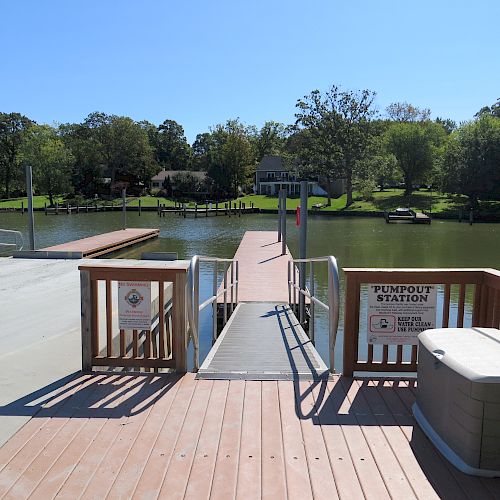 A wooden dock extends into a calm lake with a sign reading "PUMPOUT STATION" on the side railing. Residential homes and trees are visible in the background.