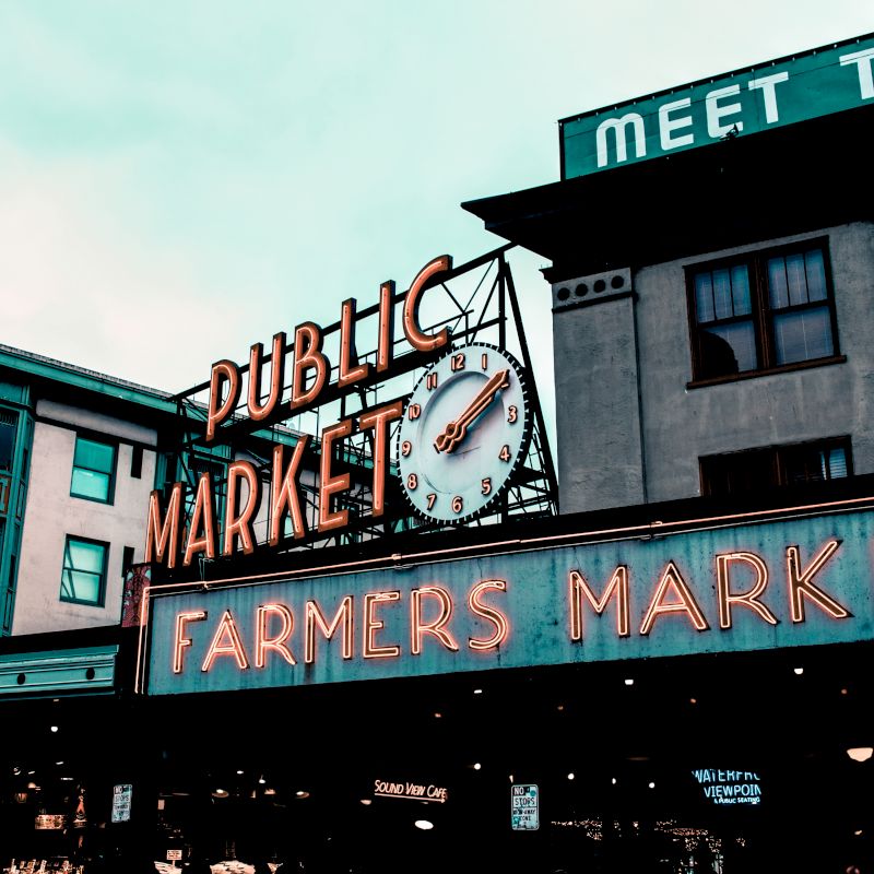 The image features the iconic "Public Market" and "Farmers Market" signs at Pike Place Market in Seattle, with surrounding buildings and a cloudy sky.
