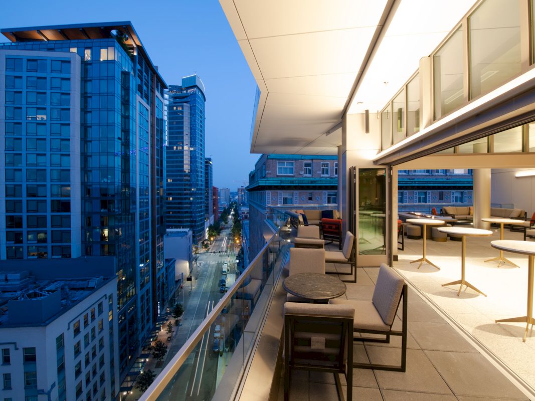 This image shows a modern balcony with seating and tables, overlooking a cityscape with tall buildings at dusk.