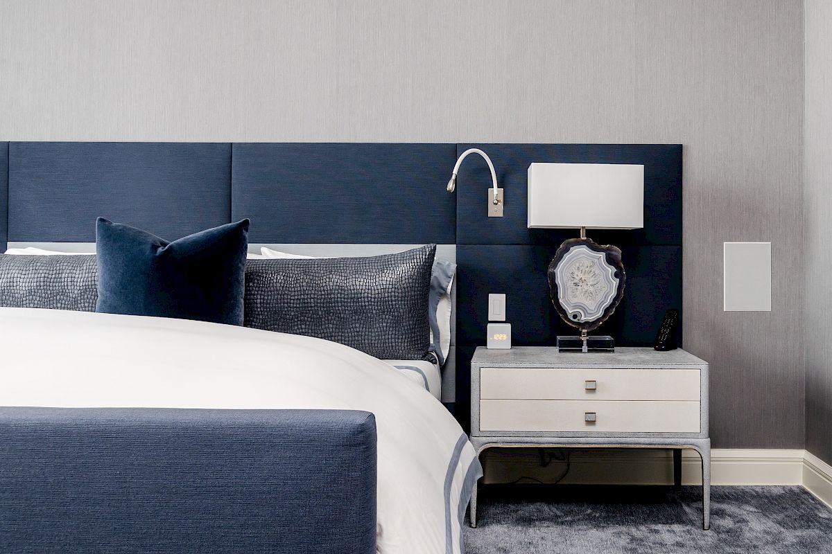 A modern bedroom features a blue and white color scheme with a neatly made bed, nightstand, lamp, and decorative elements, ending the sentence.