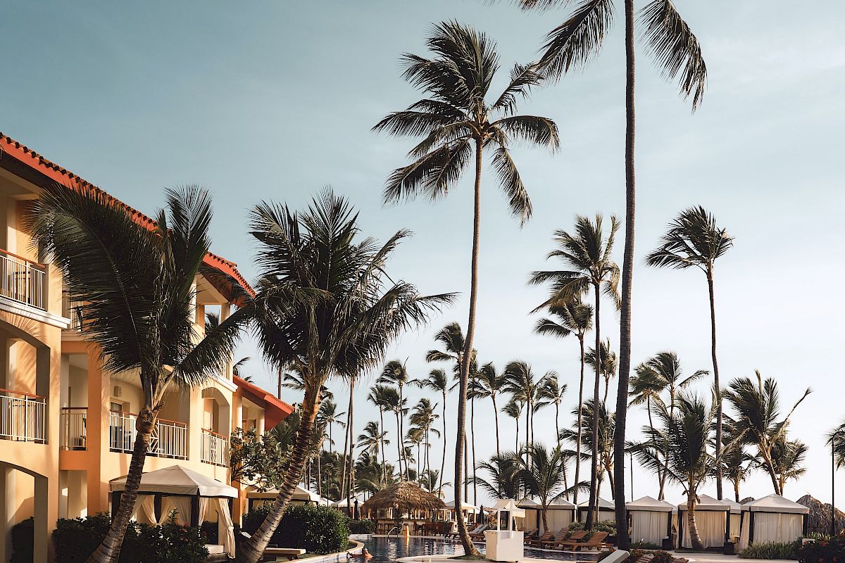 An inviting pool area with lounge chairs and tall palm trees beside a multi-story building under a clear sky, suggesting a tropical resort setting.