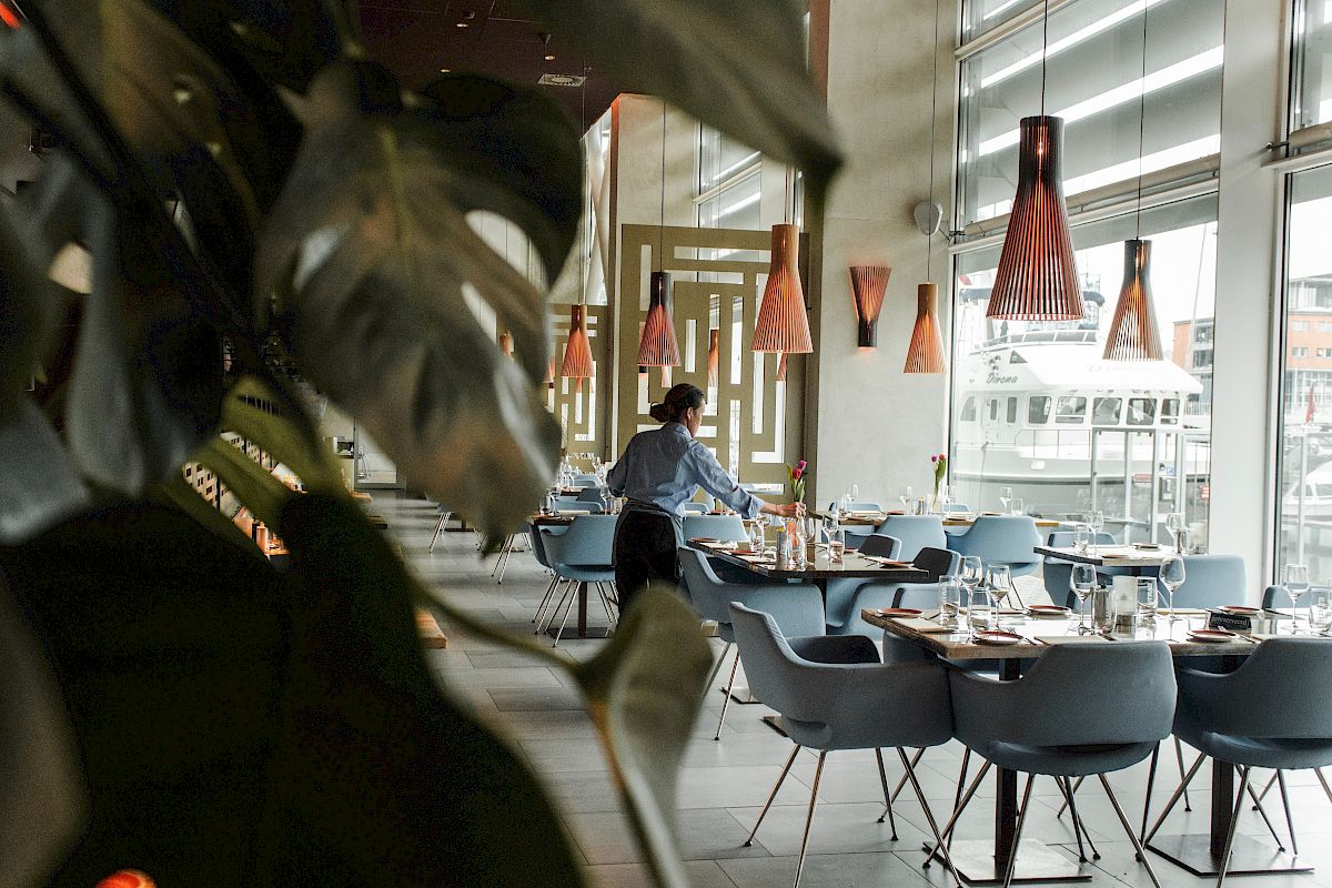 The image shows a person setting tables in a modern restaurant decorated with hanging lights and large windows, with some plants in the foreground.