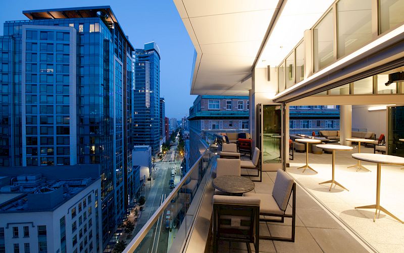 A modern cityscape at dusk showing a view from a balcony with outdoor seating, overlooking a street and tall buildings.