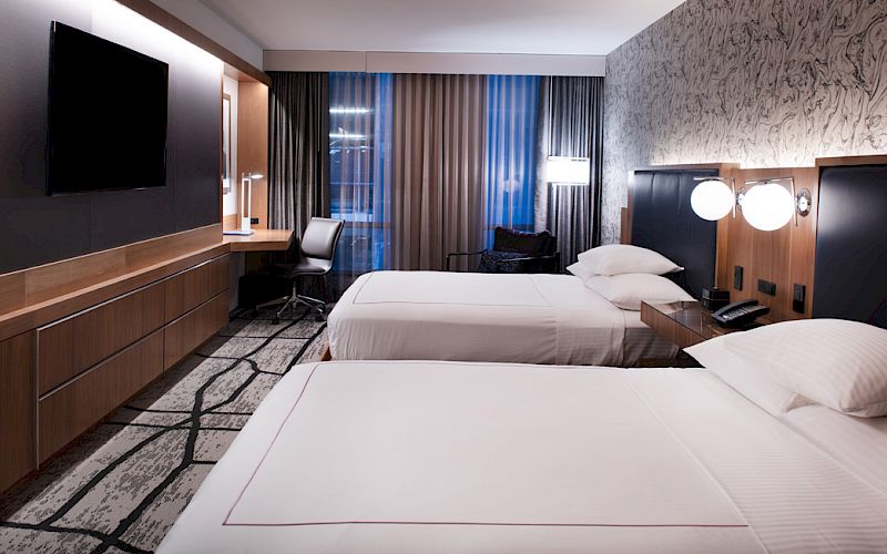 The image shows a modern hotel room with two beds, a flat-screen TV, a work desk with a chair, and large windows with curtains.